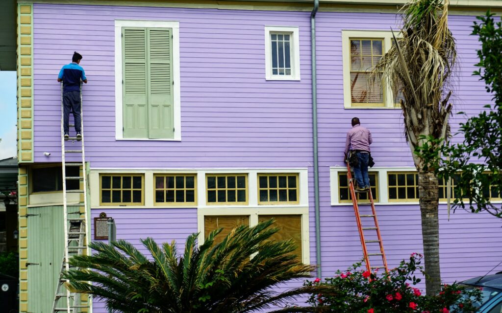 Two workers on ladders cleaning and prepping the siding of a two-story house