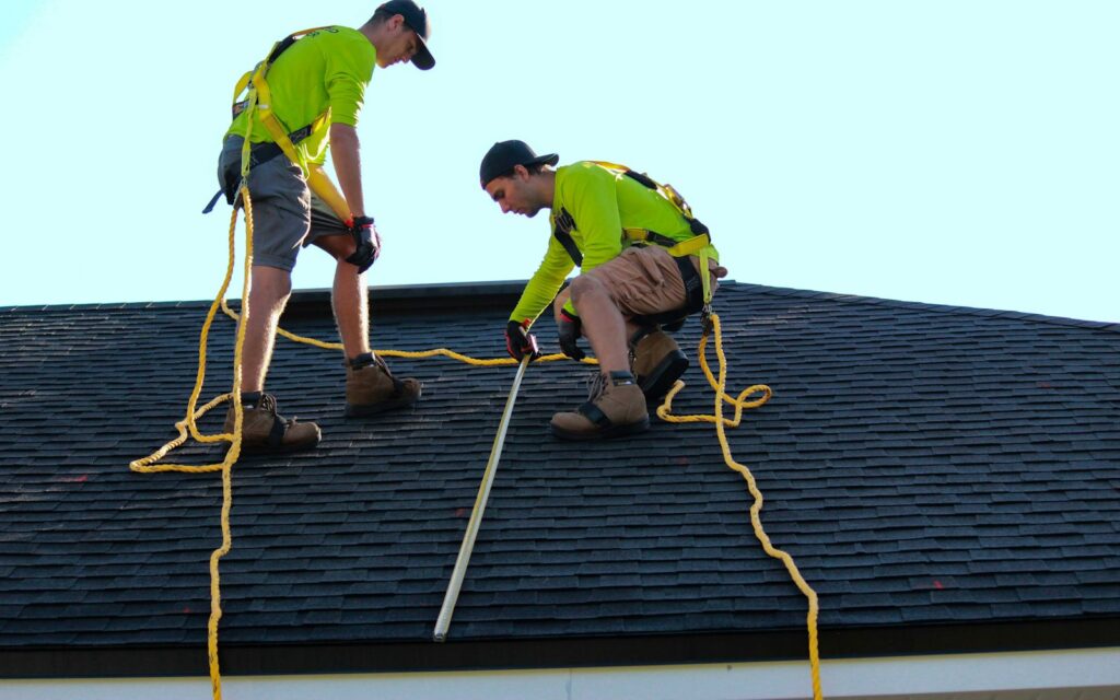 Two workers on an asphalt shingle roof using safety ropes